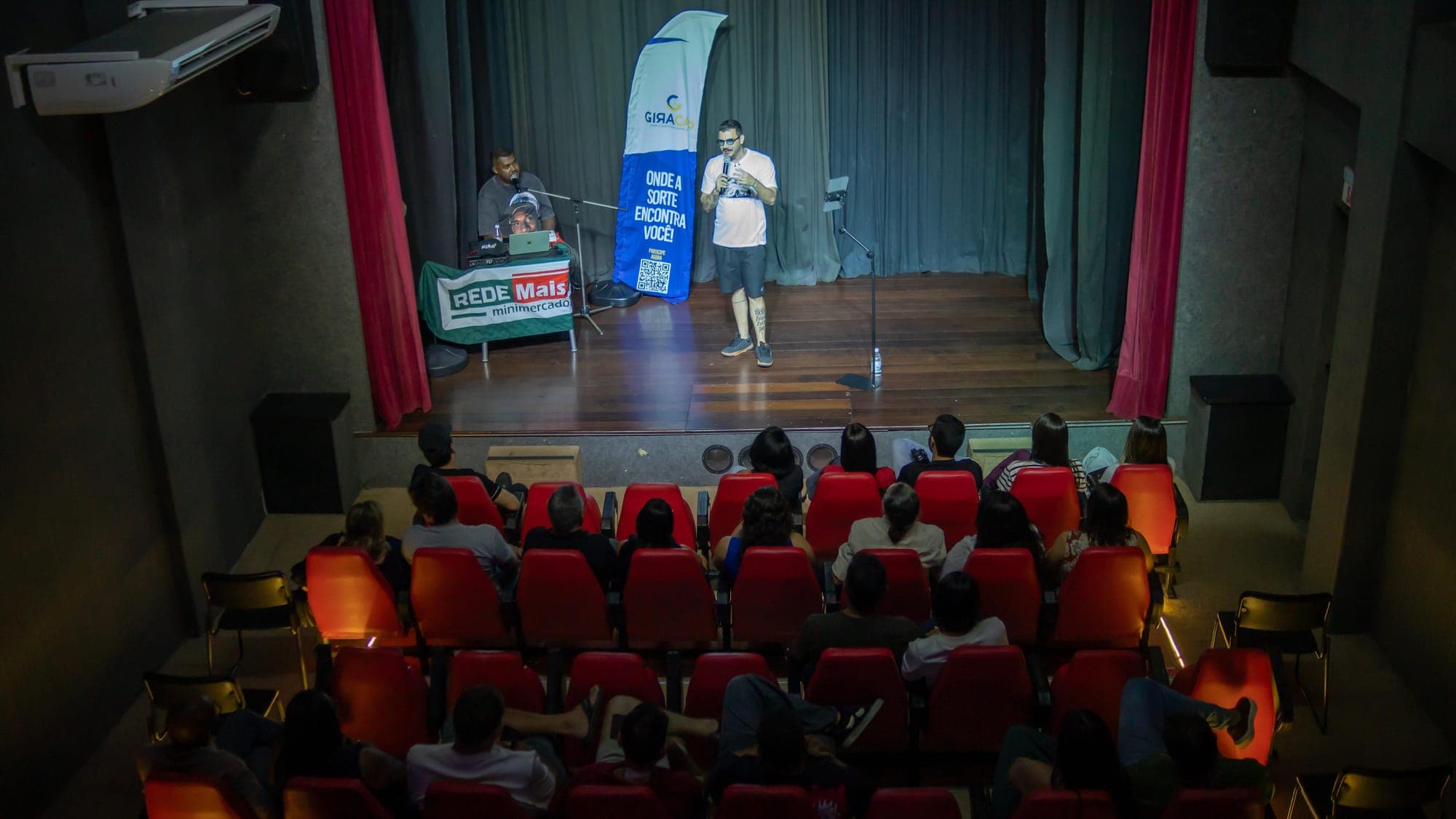 Palco do teatro visto de cima, com Igor em cena e a plateia sentada nas poltronas vermelhas